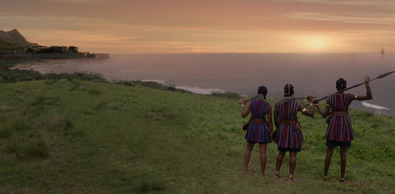 Three women part of the legendary all-woman African army carry their weapons, machetes and a spear while looking over the setting sun on the ocean.